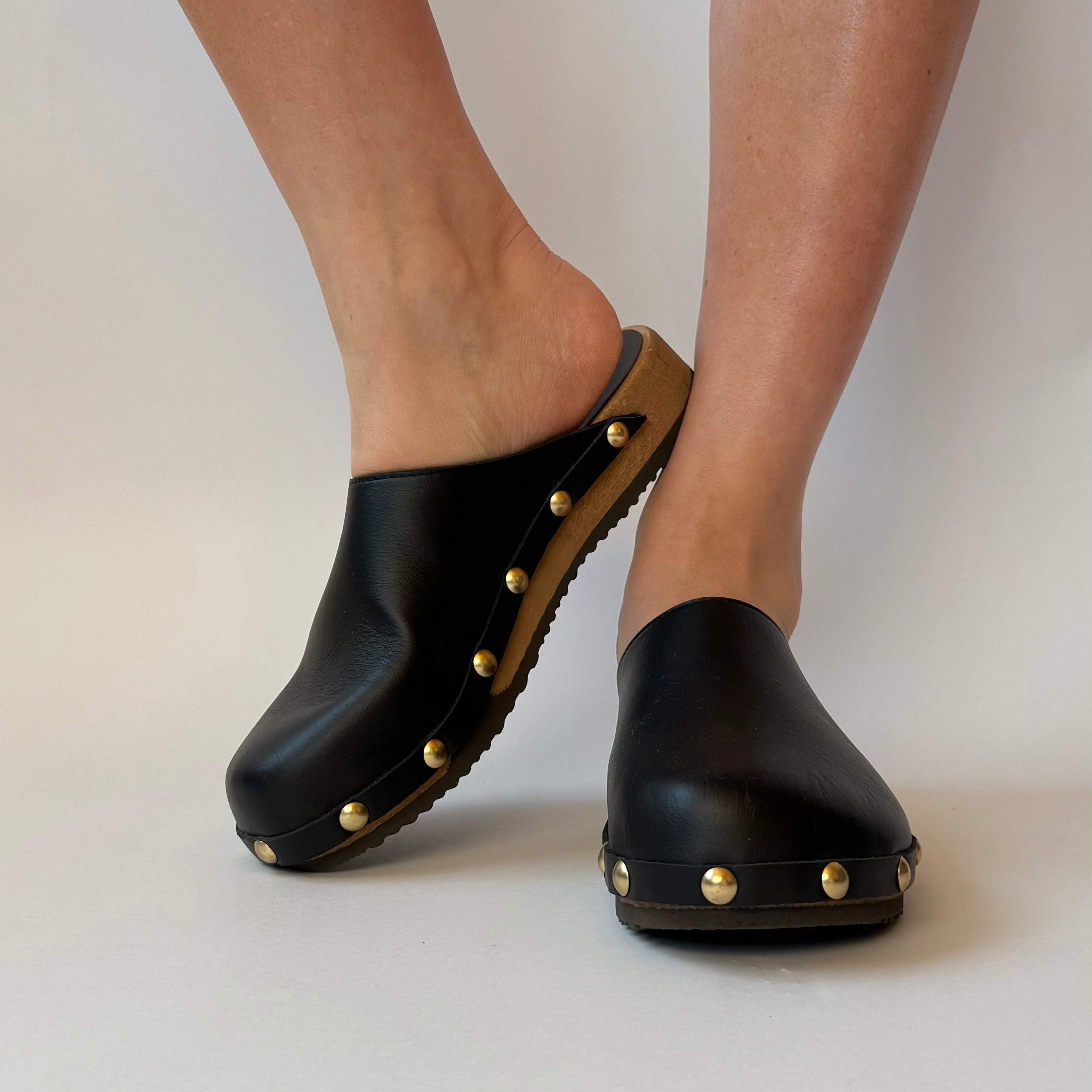 Black clogs with gold studs worn by a person on a plain background