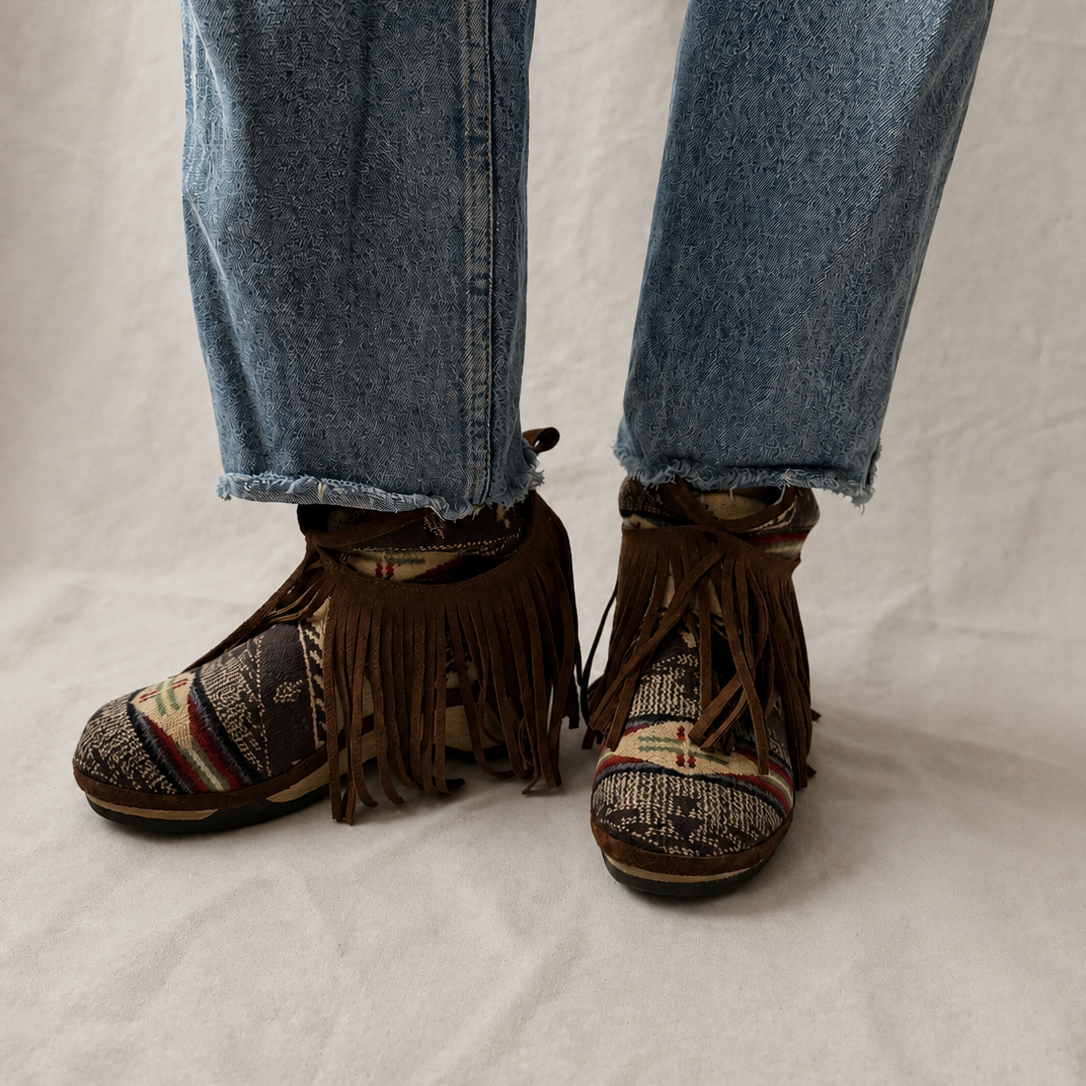 A person wearing fringed brown clog boots with patterned woven uppers, paired with light blue jeans, standing against a neutral off-white fabric backdrop.