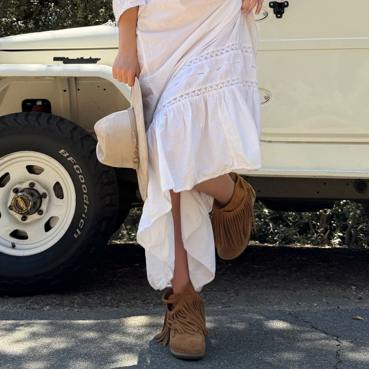 A woman in a white dress and KUNSTEN Holly fringe boots beside a cream vintage car in California
