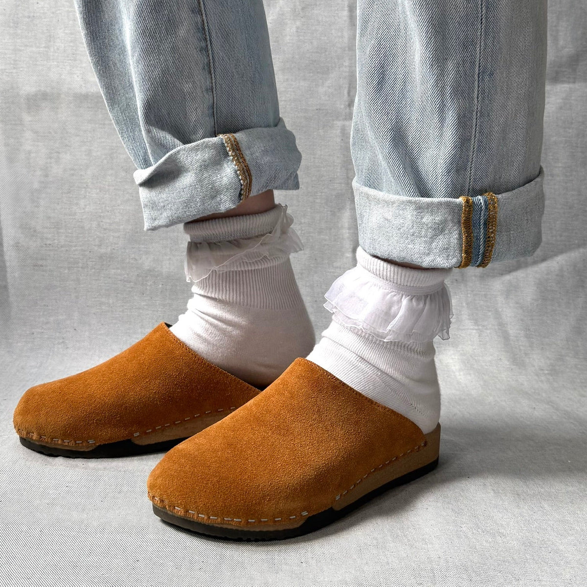 Brown suede slippers worn with light blue jeans and white socks on a neutral background