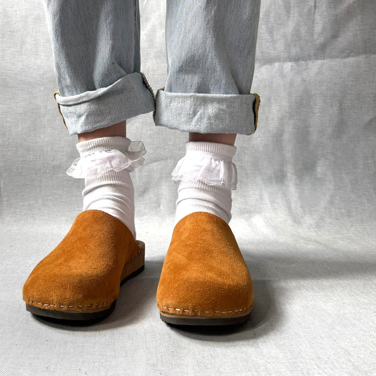 Person wearing brown suede shoes, light blue jeans, and white socks with ruffles on a plain background