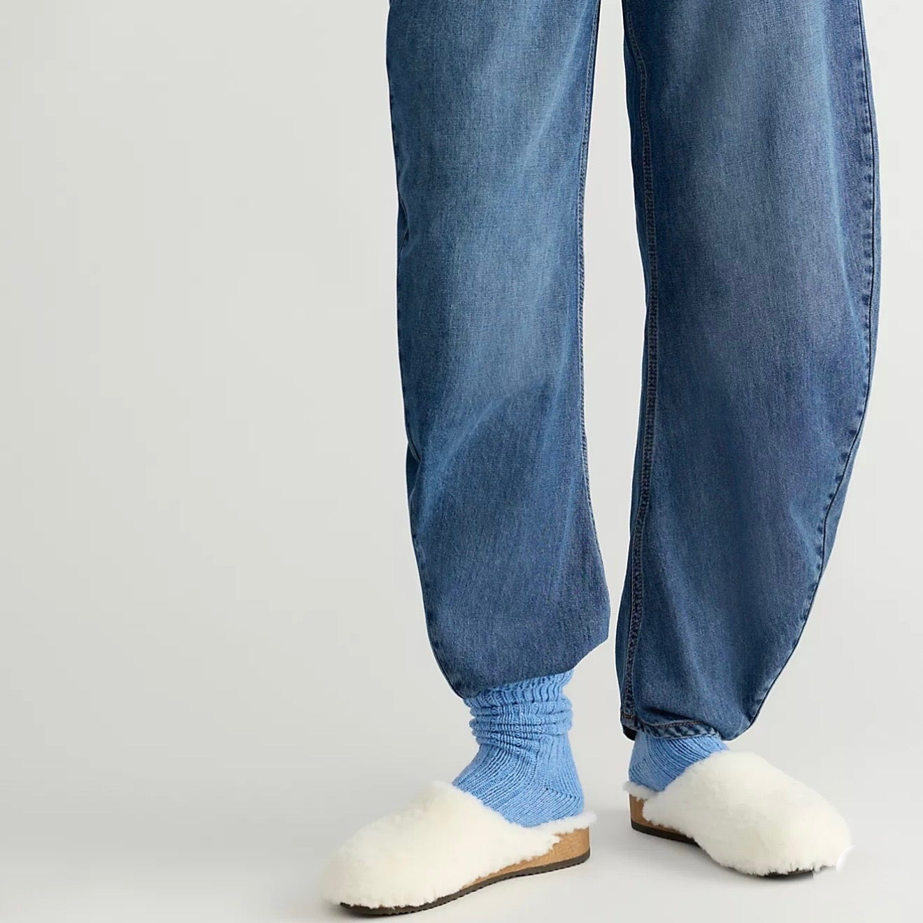 A person wearing loose blue denim jeans with gathered cuffs, paired with light blue socks and white fluffy slippers, photographed against a plain light gray studio background.