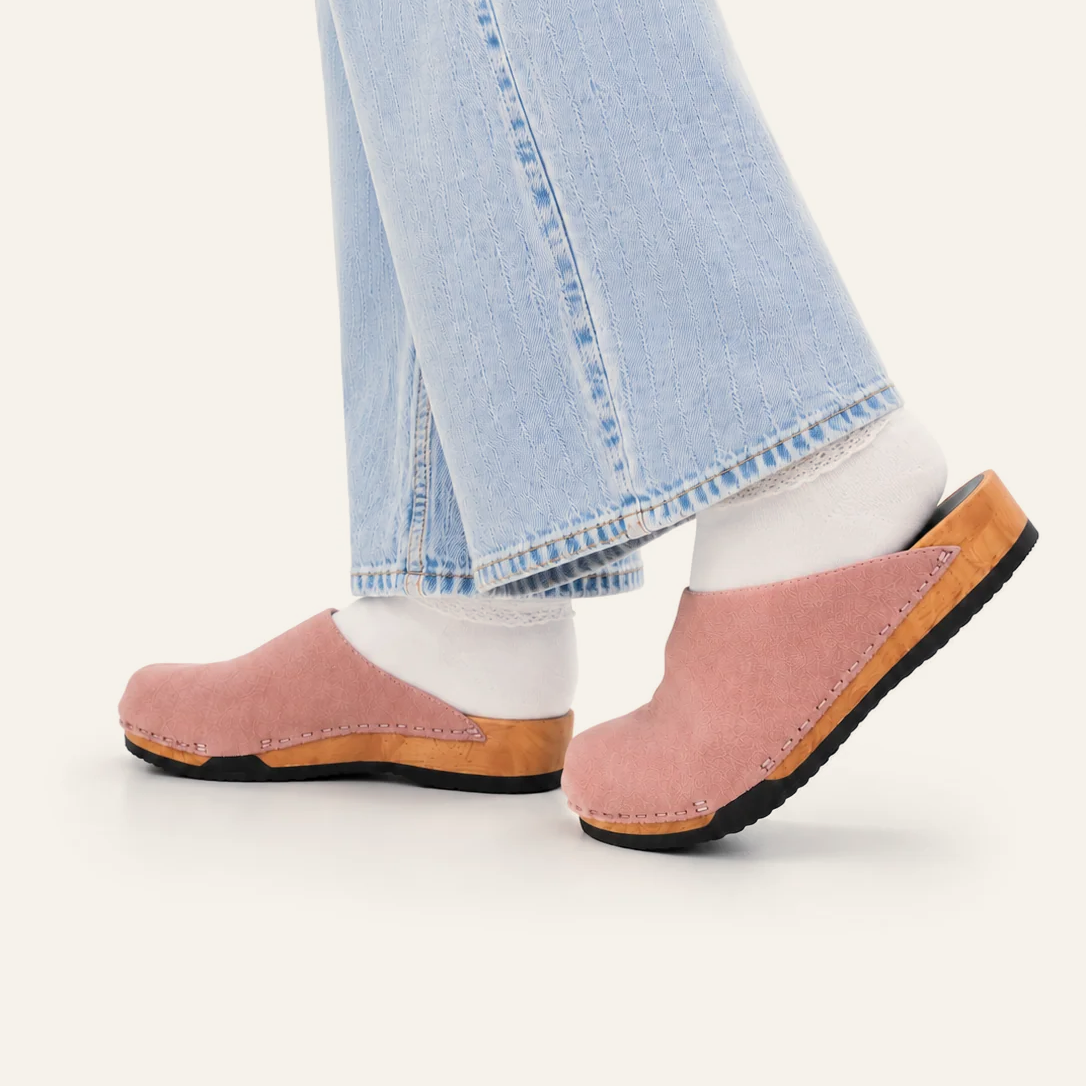 A person wearing light blue jeans, white socks, and dusty pink clogs with tan soles, shown from the calves down against a plain off-white background.
