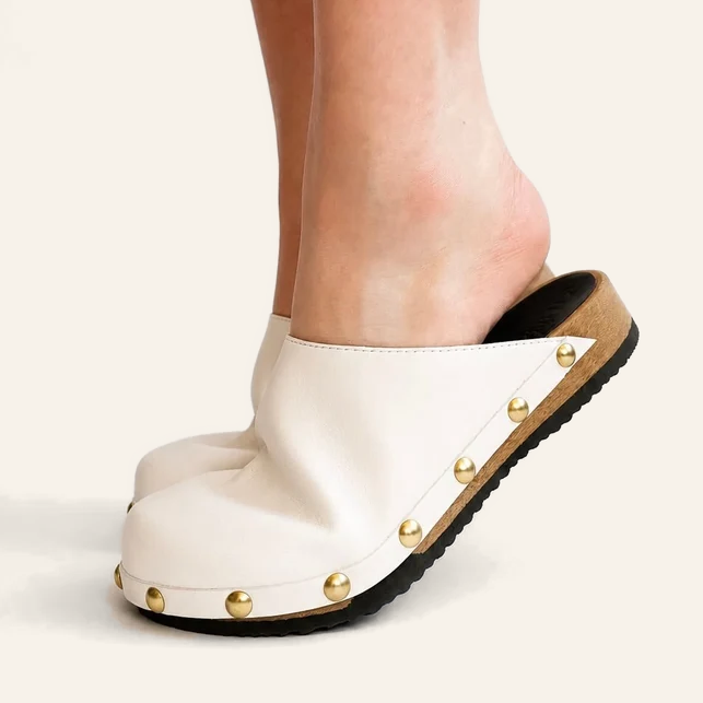 A close-up of a person's foot wearing white leather comfort clogs with gold-tone studs and a black sole, shown against a plain light beige background.