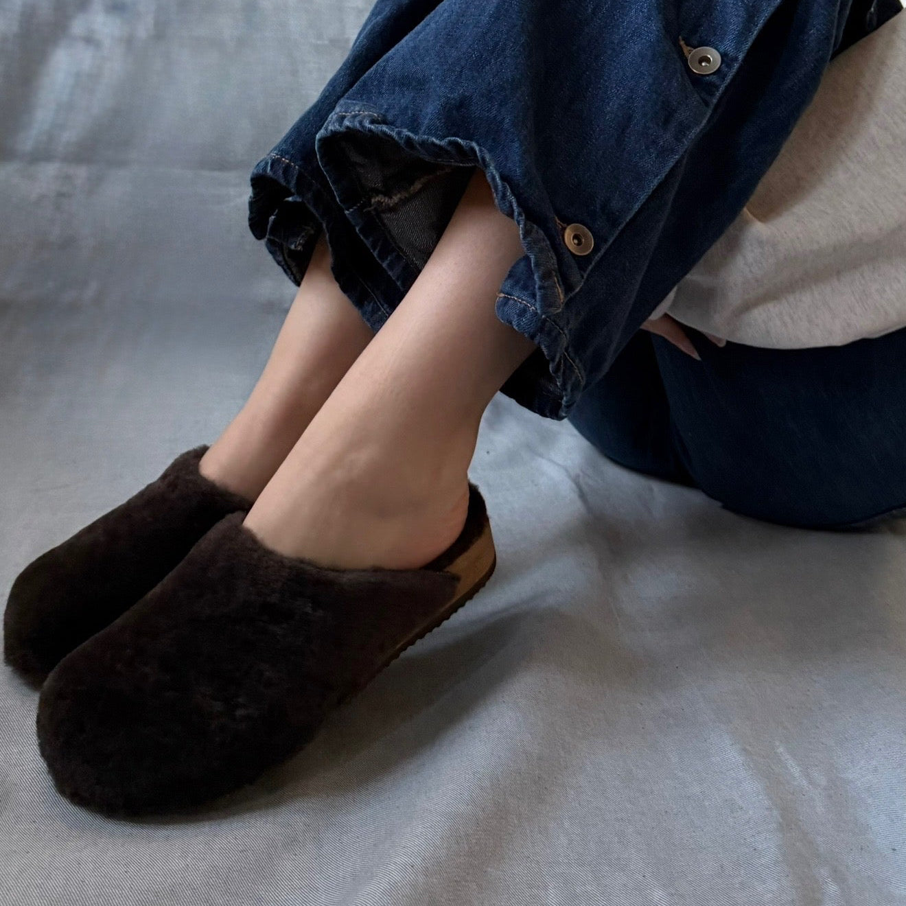A person wearing dark brown fuzzy slip-on slippers with a light tan lining, paired with dark blue denim pants and a white top, sitting on a light gray fabric surface.