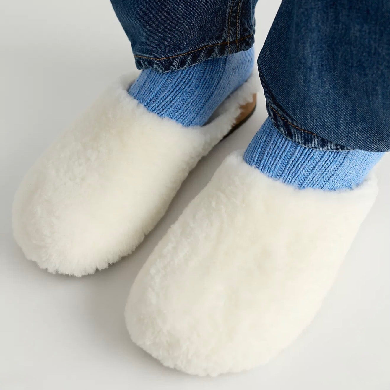White fluffy slip-on slippers worn with light blue ribbed socks and dark blue denim jeans, shown from a top-down angle on a light gray background.