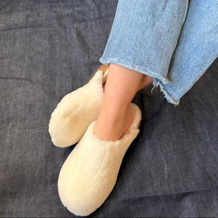 A person wearing cream-colored fuzzy slip-on slippers with light blue denim jeans, sitting on a dark gray textured fabric surface.