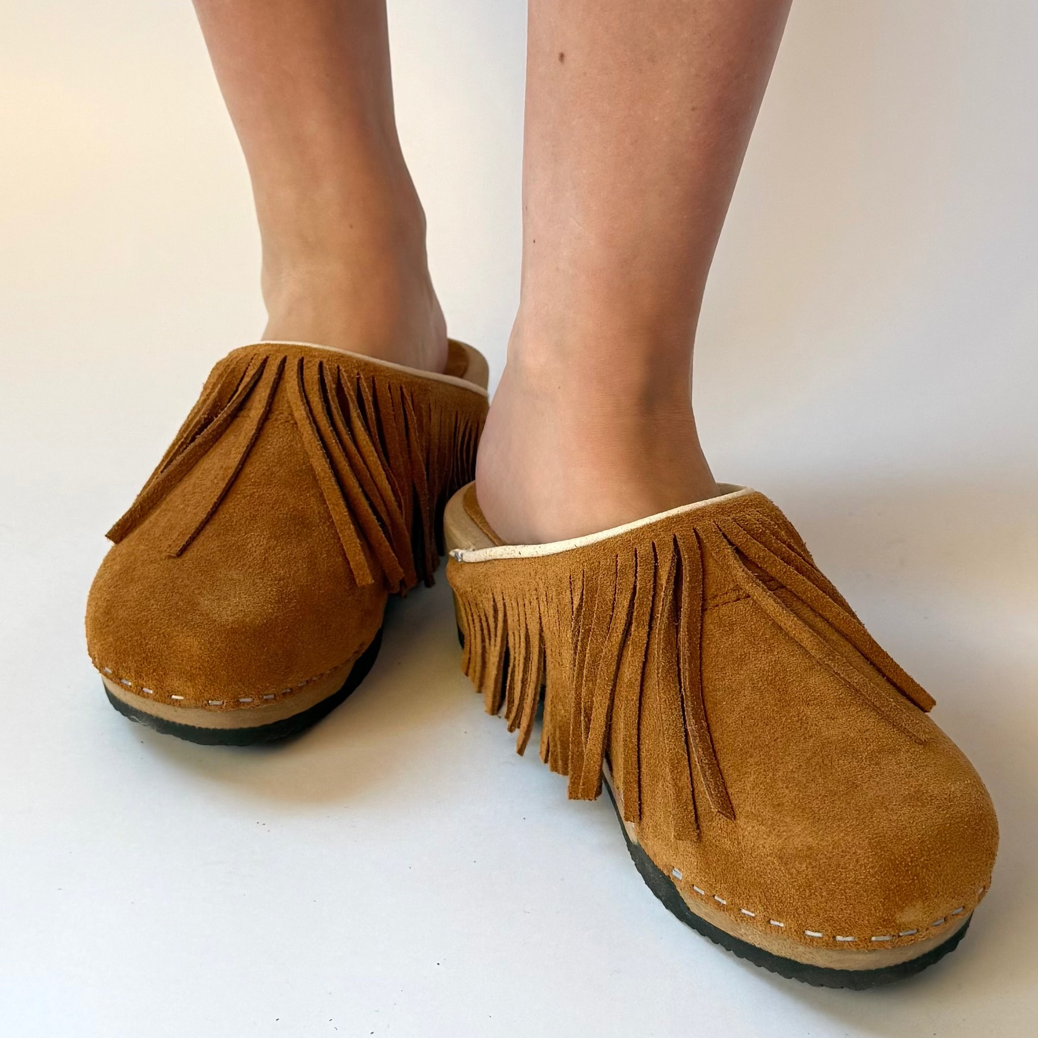 A pair of tan suede fringe clogs worn on feet, shown from the side against a plain white background.