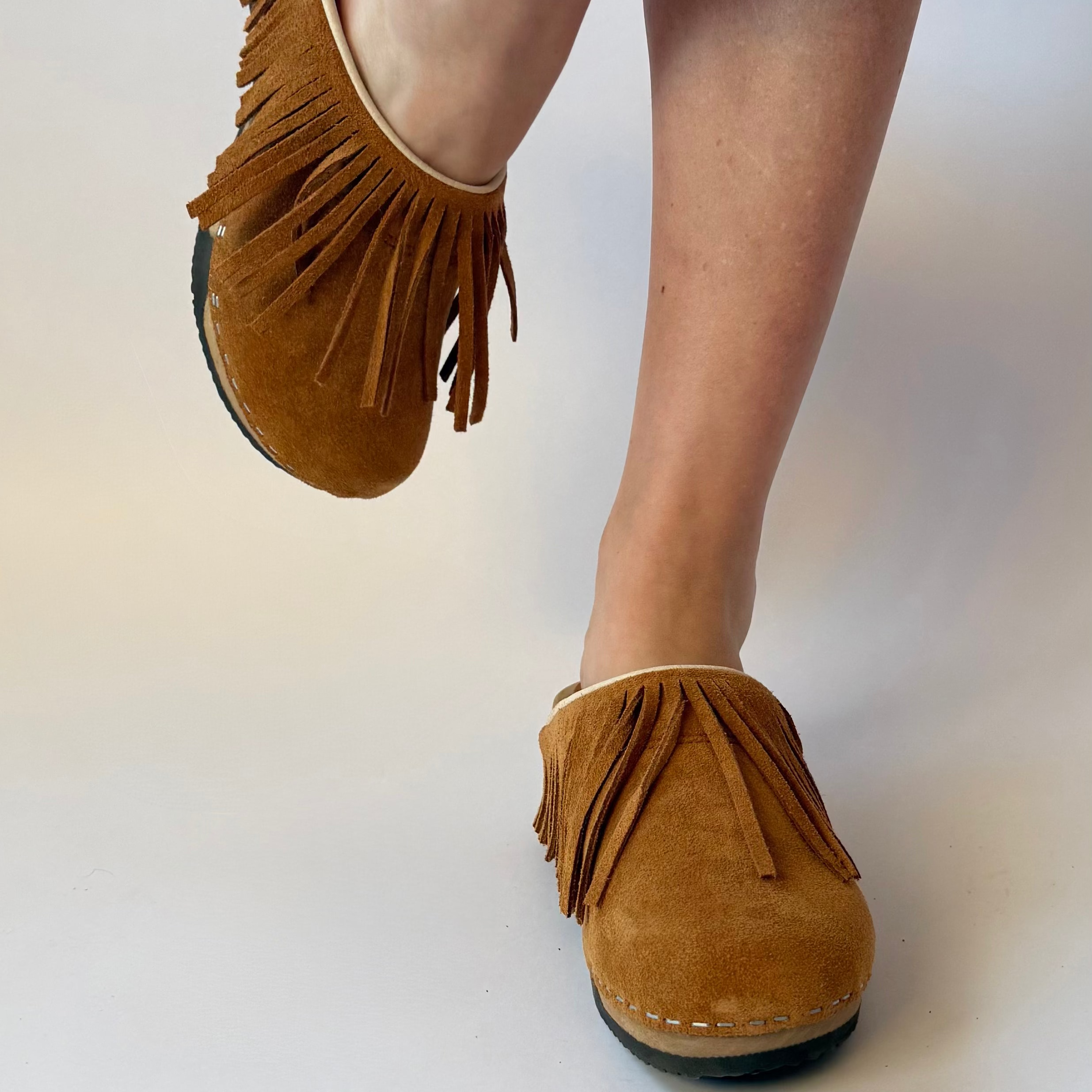 A pair of tan suede clogs with fringe detailing, shown on a model's feet against a plain light background.