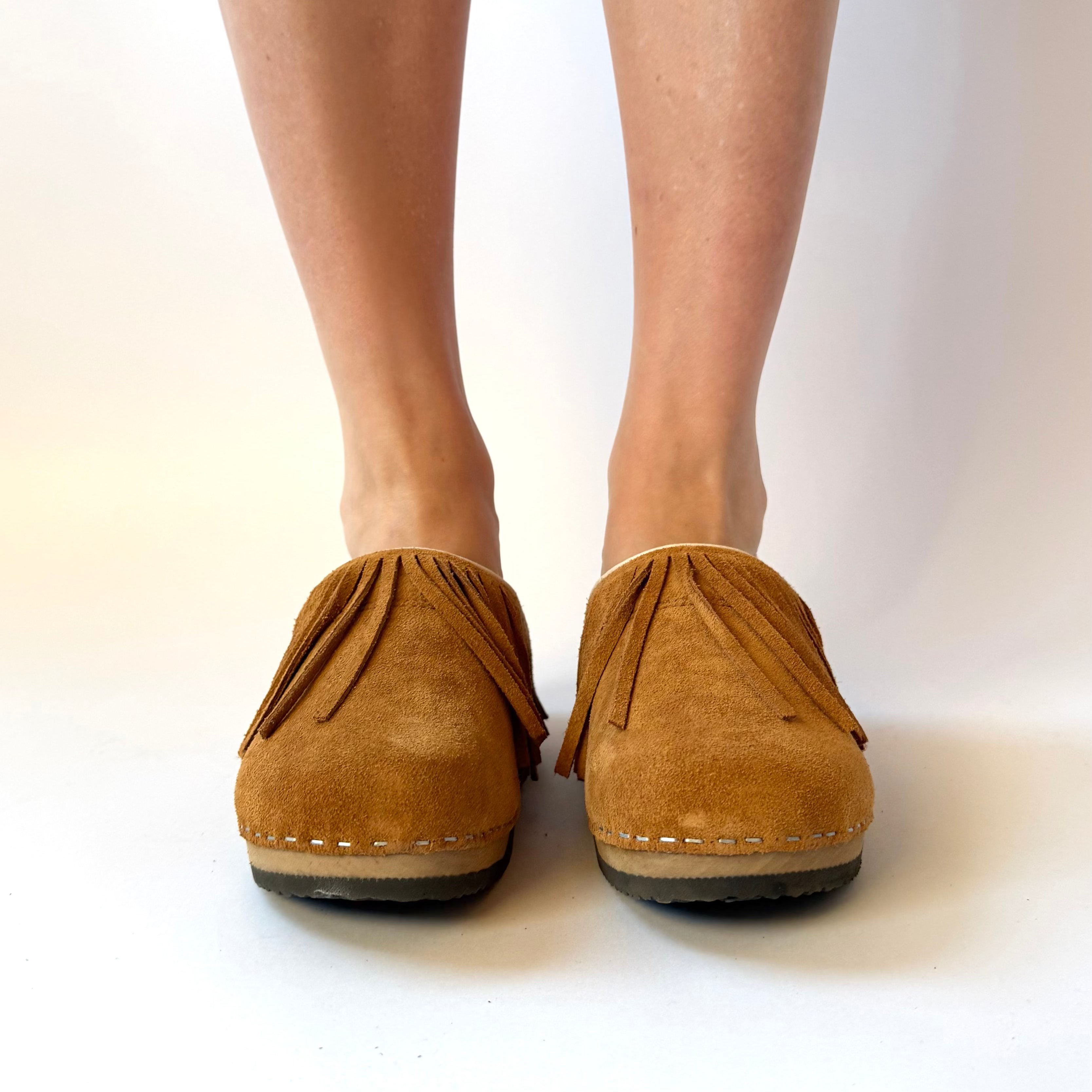 A pair of tan suede clogs with fringe detailing, shown on a person's feet against a plain white studio background.