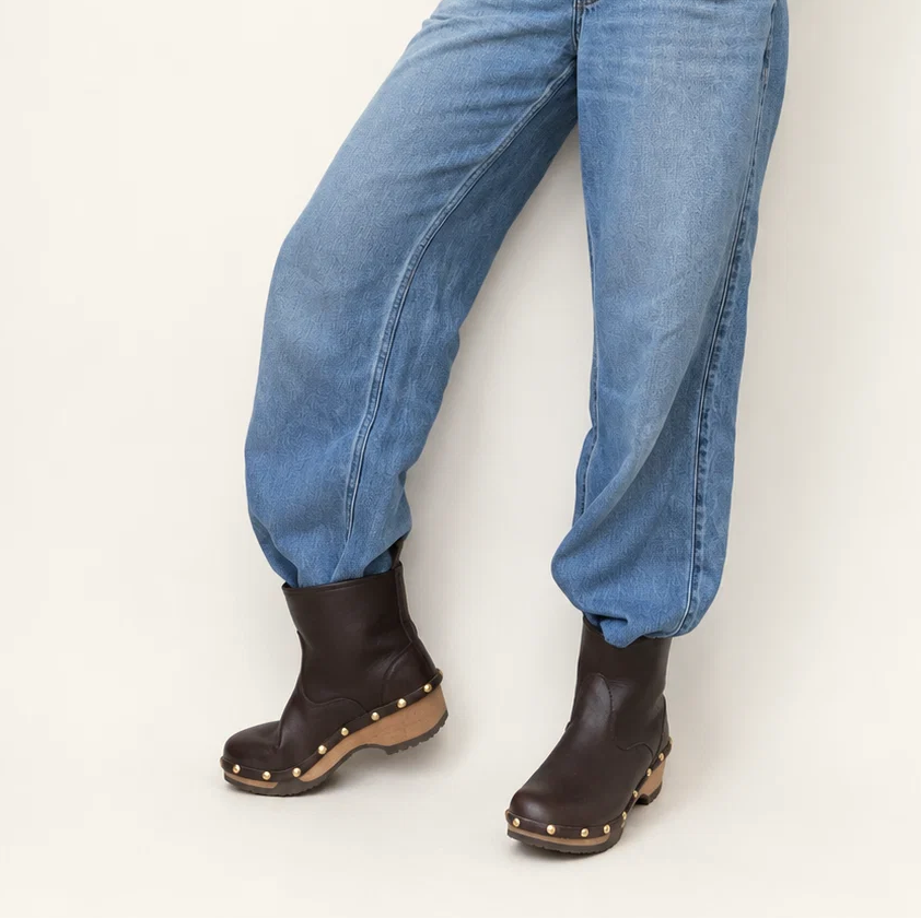 A person wearing light blue relaxed-fit jeans and dark brown leather boots with gold-tone studs, shown from the waist down against a plain off-white studio background.