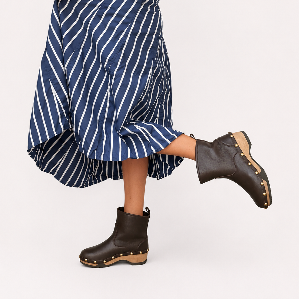 A woman wearing a navy blue and white vertically striped midi skirt, a white sleeveless top, and dark brown leather ankle boots with gold studs, captured against a solid off-white studio background.