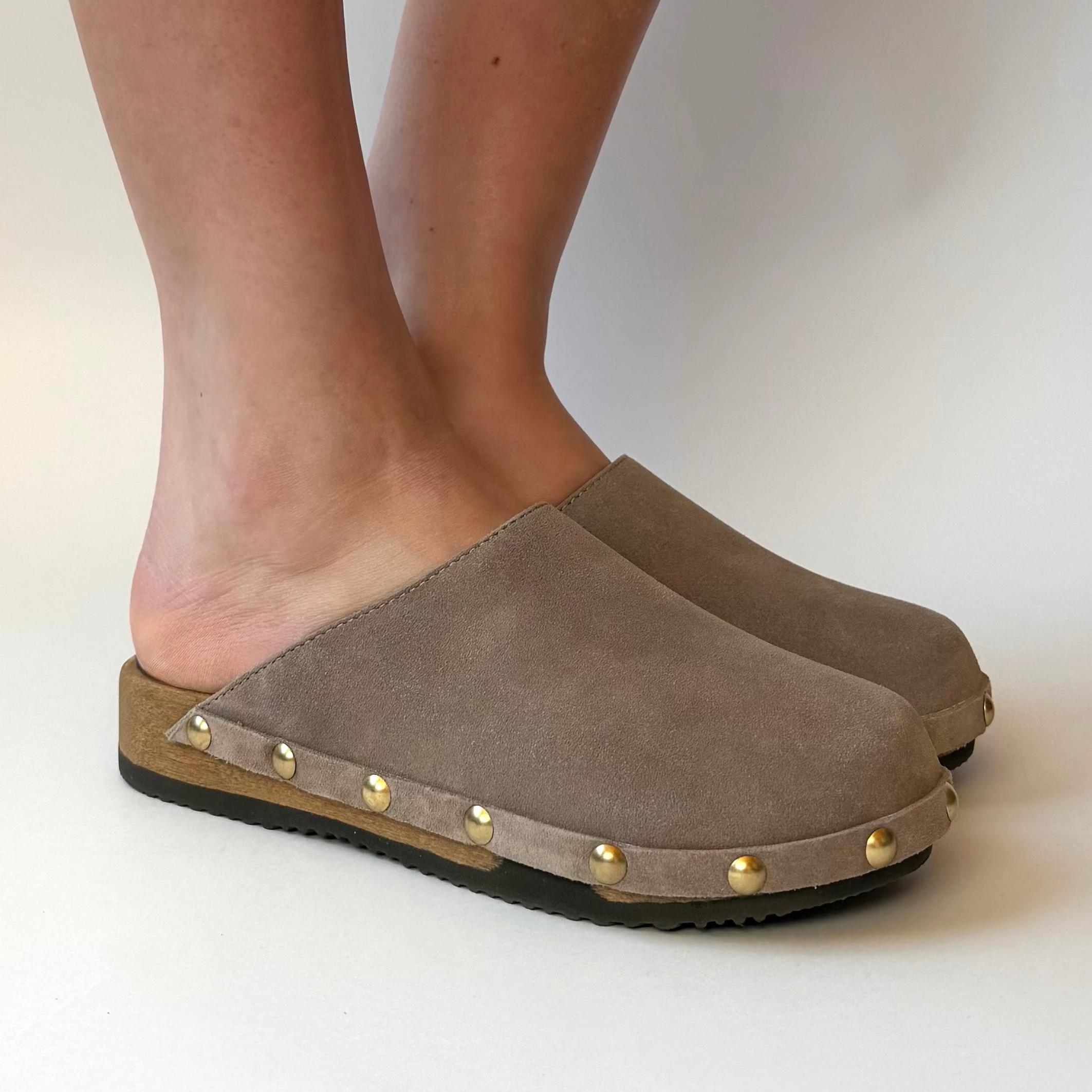 Brown suede clogs with gold studs worn by a person on a white background