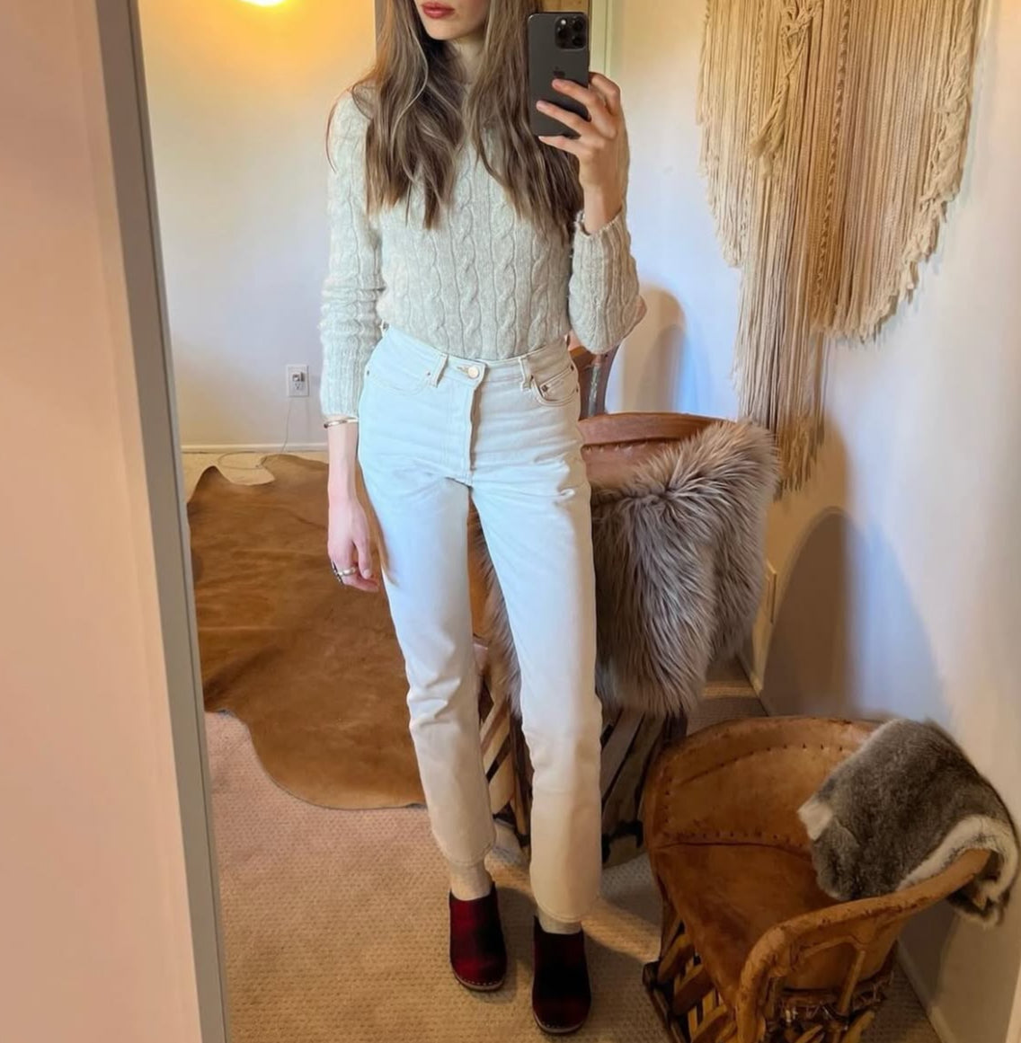Person taking a mirror selfie wearing a light sweater and white pants in a room with a chair and rug.