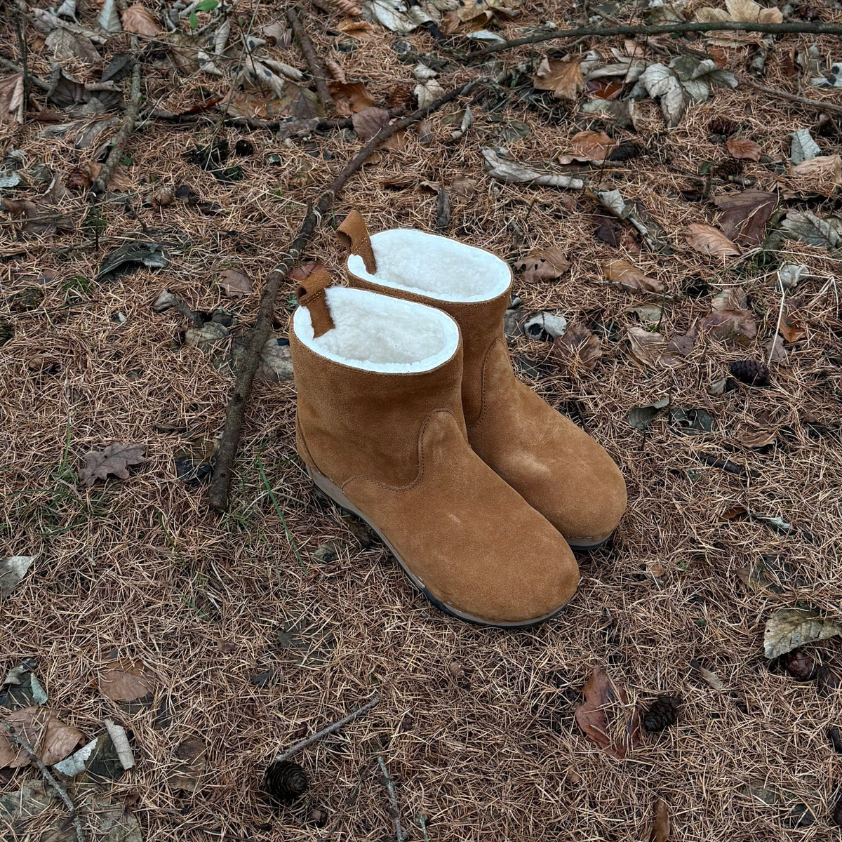 Pair of brown suede boots with white fur lining on a ground covered with dry leaves and twigs.