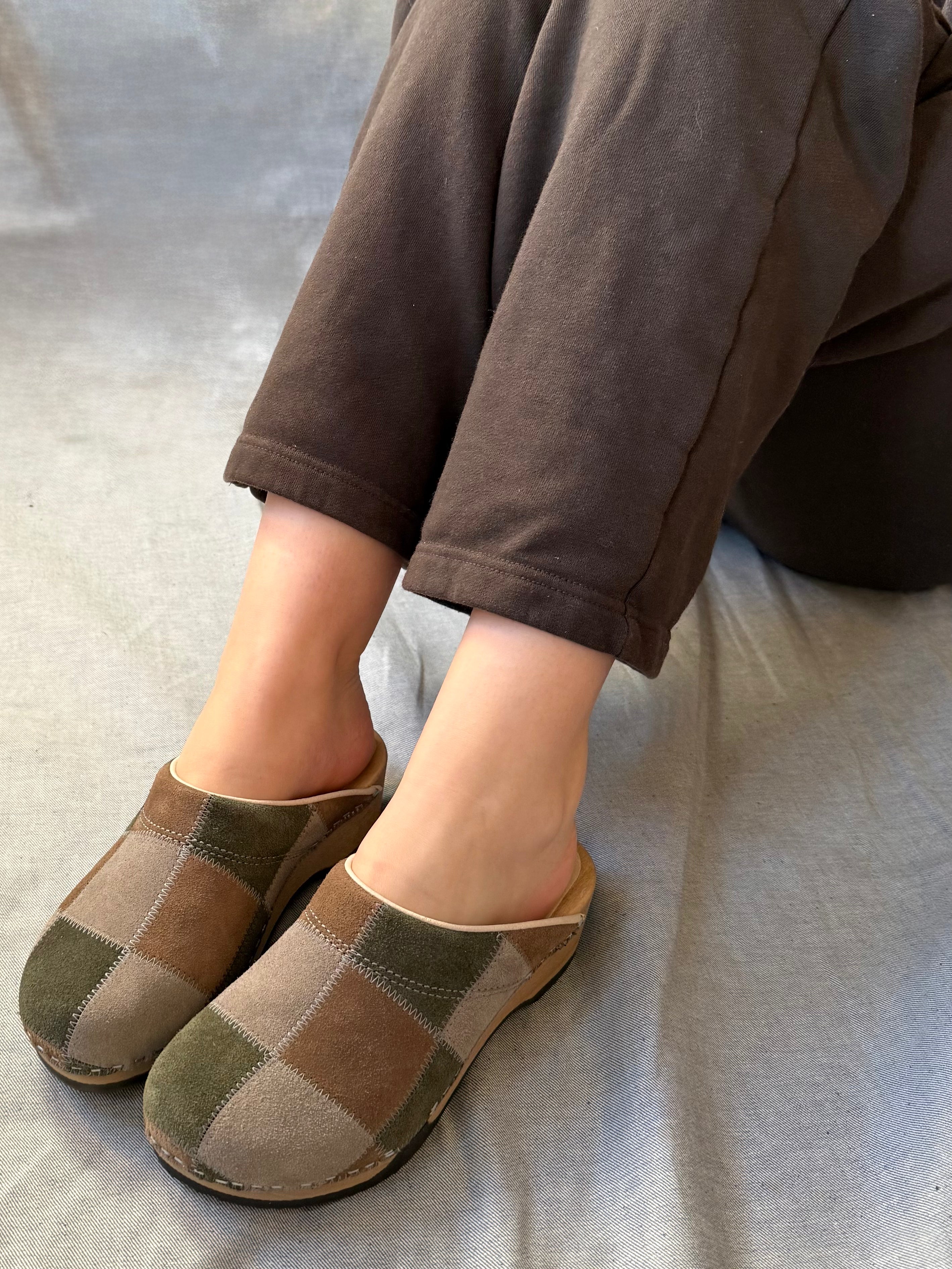 Pair of checkered clogs worn with brown pants on a light gray surface.