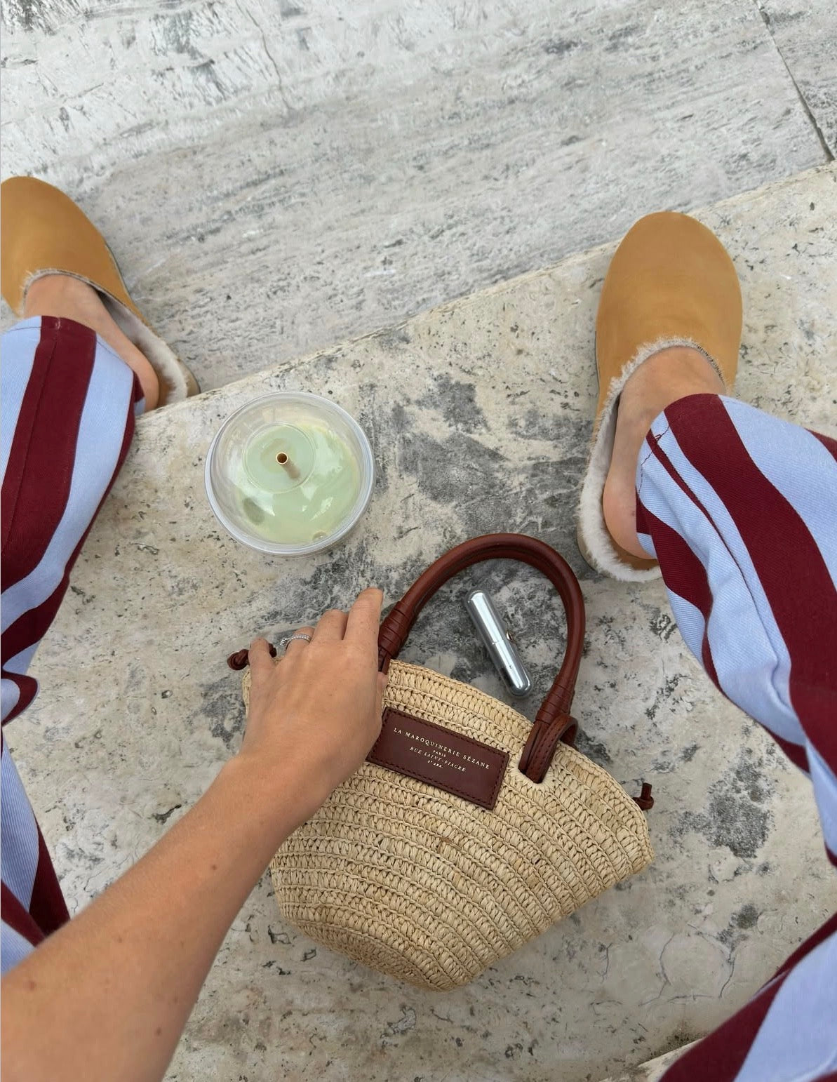 Person holding a straw handbag on a stone surface with a drink and a phone.