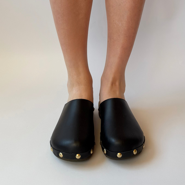 Black clogs with gold studs worn by a person on a white background