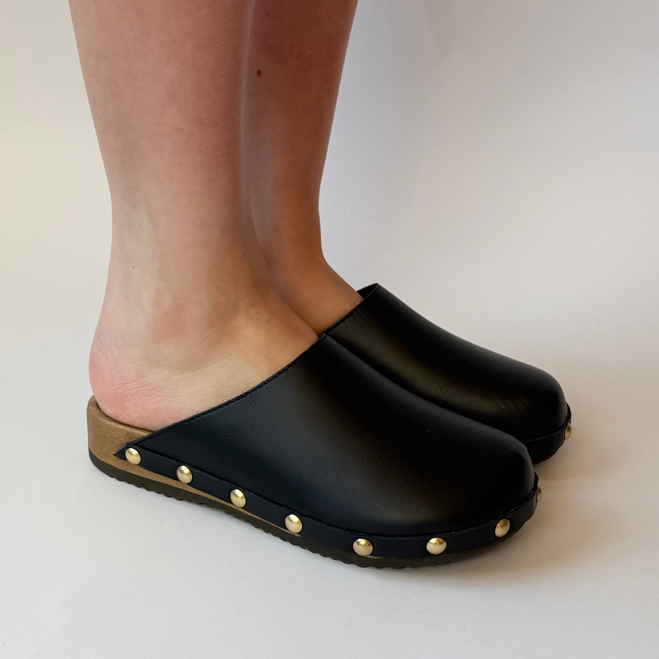 Black clogs with gold studs worn by a person on a plain background