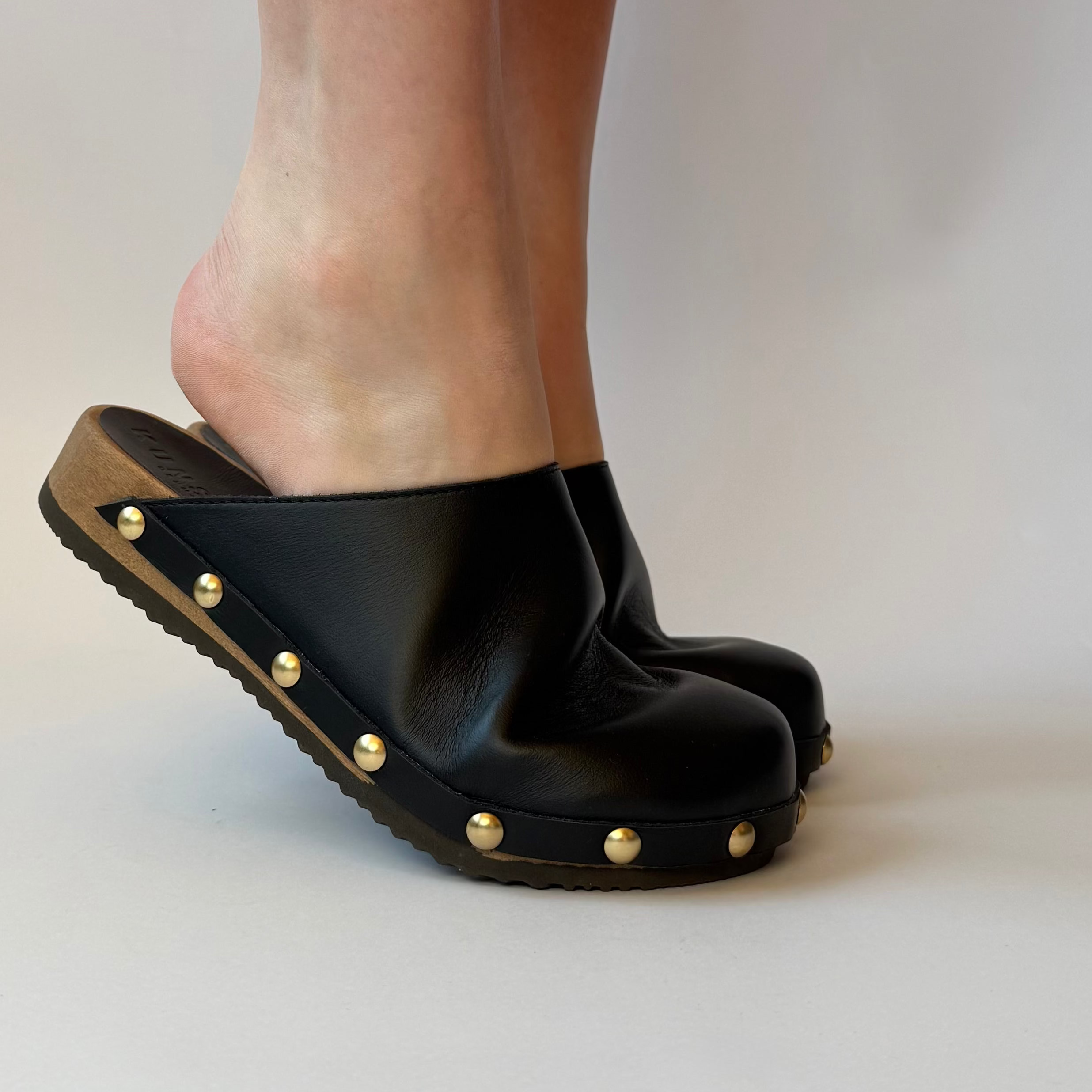 Black clogs with gold studs worn by a person on a neutral background