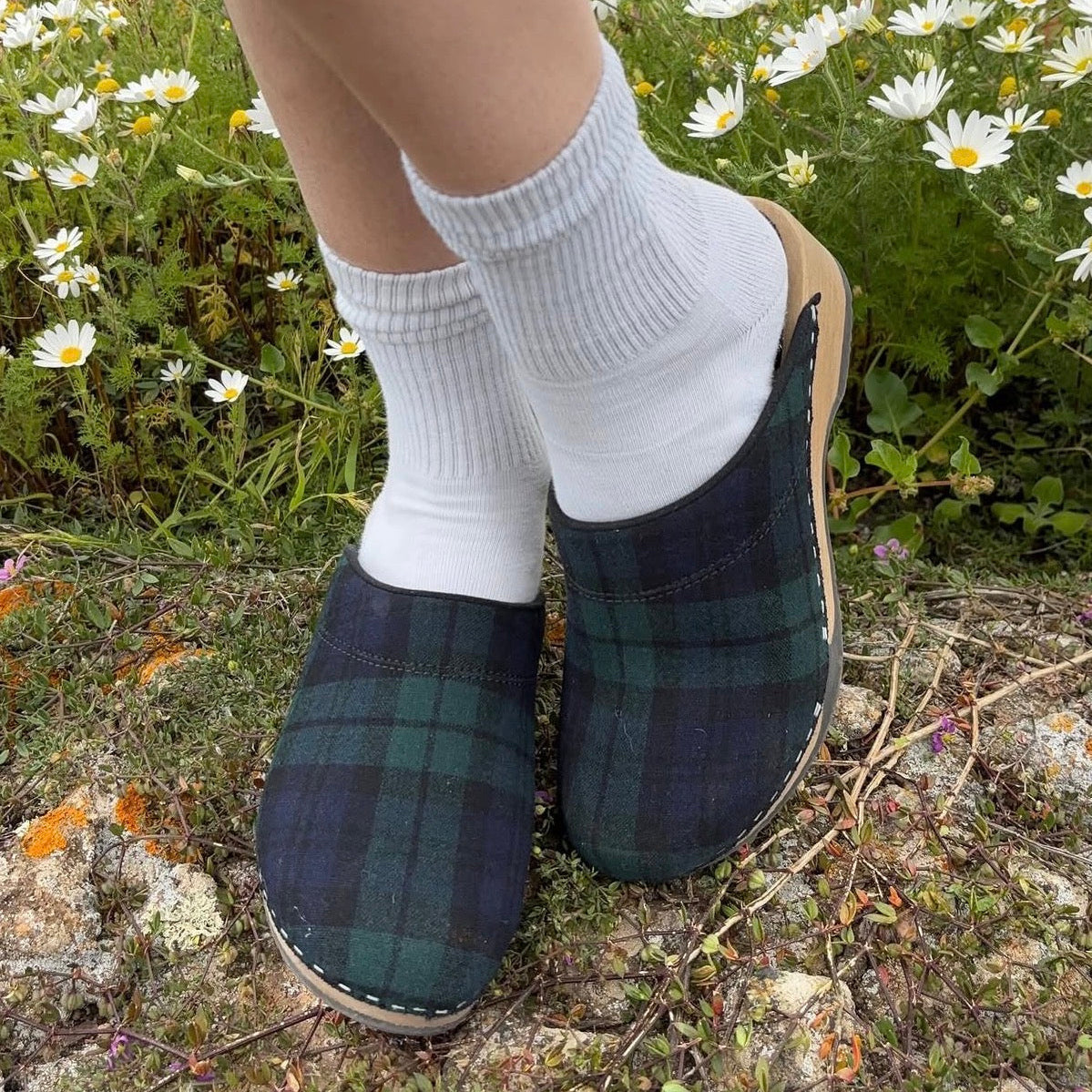 Plaid clogs worn with white socks on a natural background with flowers.