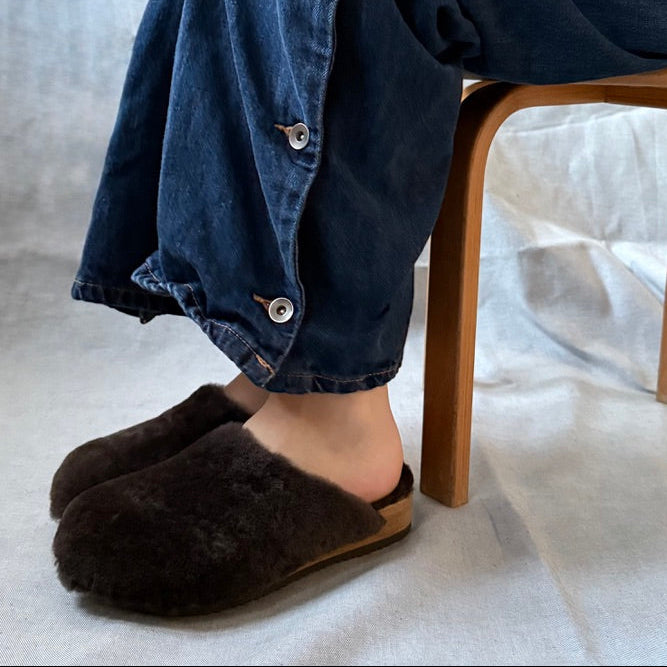 A person seated on a wooden stool wearing loose dark blue denim pants with silver buttons, a light beige sweater, and dark brown fuzzy slip-on slippers against a soft light gray backdrop.