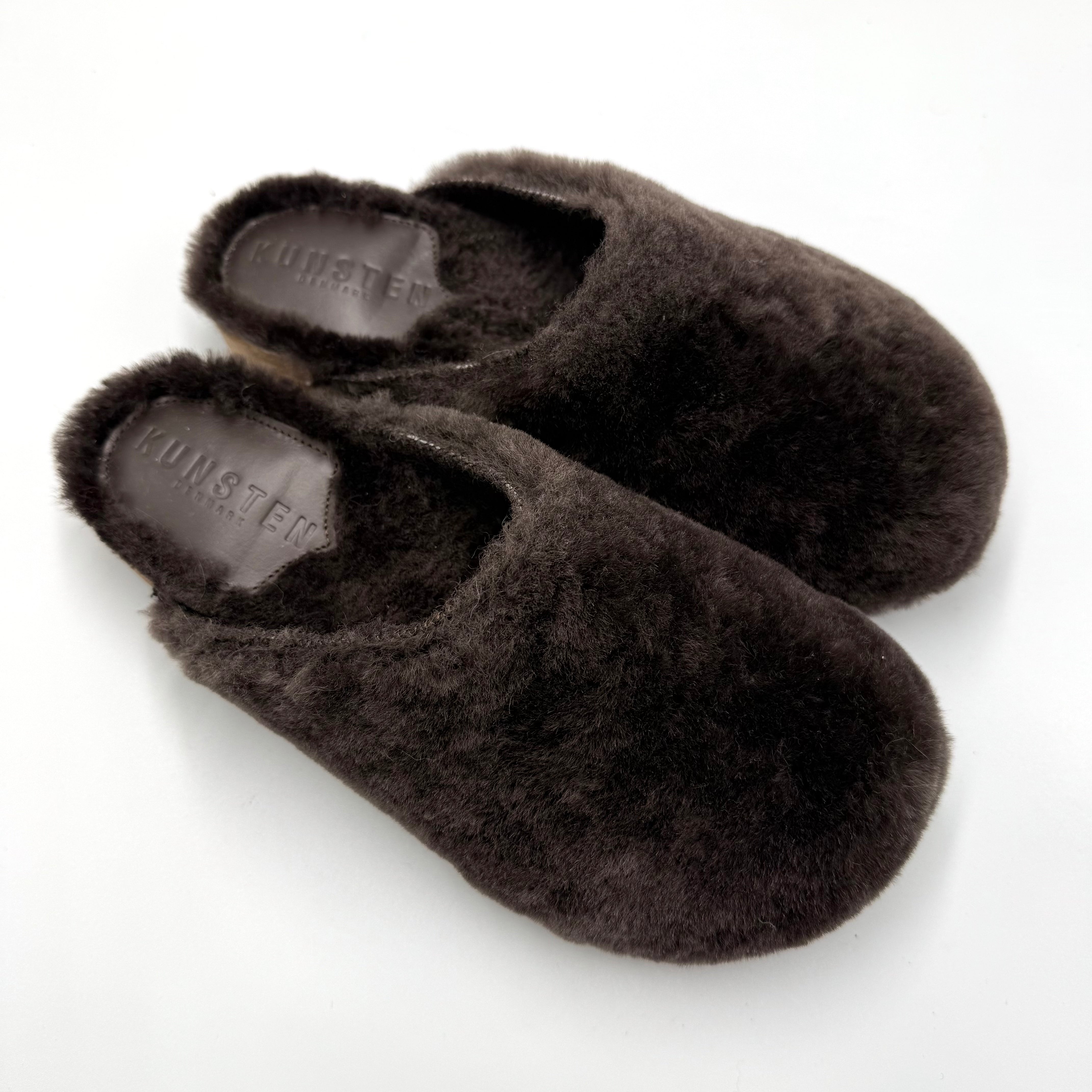 A pair of dark brown slip-on slippers with a visible 'KUNSTEN' brand insole, displayed on a clean white background.