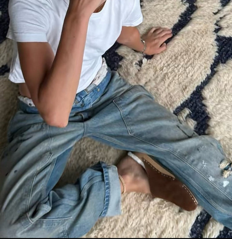 A young woman with blonde hair in a bun and glasses sits on a textured beige and navy rug, wearing a white t-shirt, light blue distressed jeans and a pair of KUNSTEN shearling clogs. 