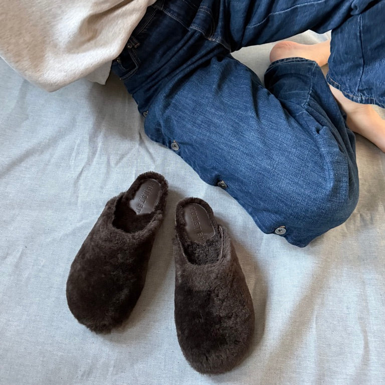 A pair of dark brown fuzzy slip-on slippers placed on a light gray fabric surface, with a person wearing blue jeans and a cream sweatshirt partially visible in the frame.