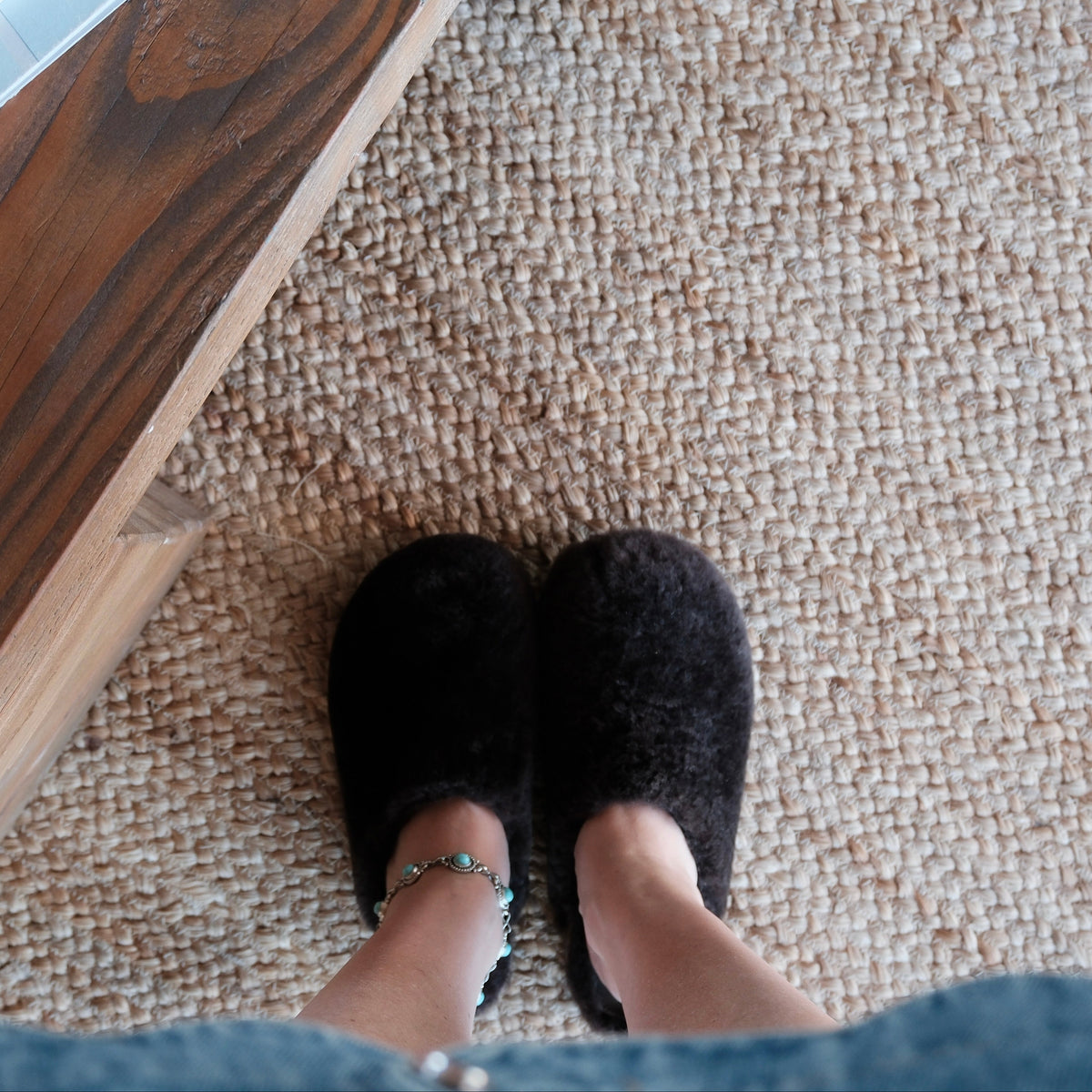 A top-down view of a person wearing a pair of brown fuzzy clogs on a textured beige carpet, with a wooden table edge visible on the left and denim fabric at the bottom.