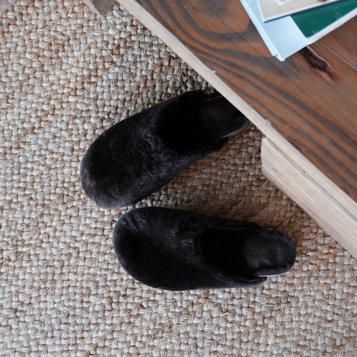A pair of brown fuzzy clogs placed on a textured beige woven rug next to a wooden table corner.