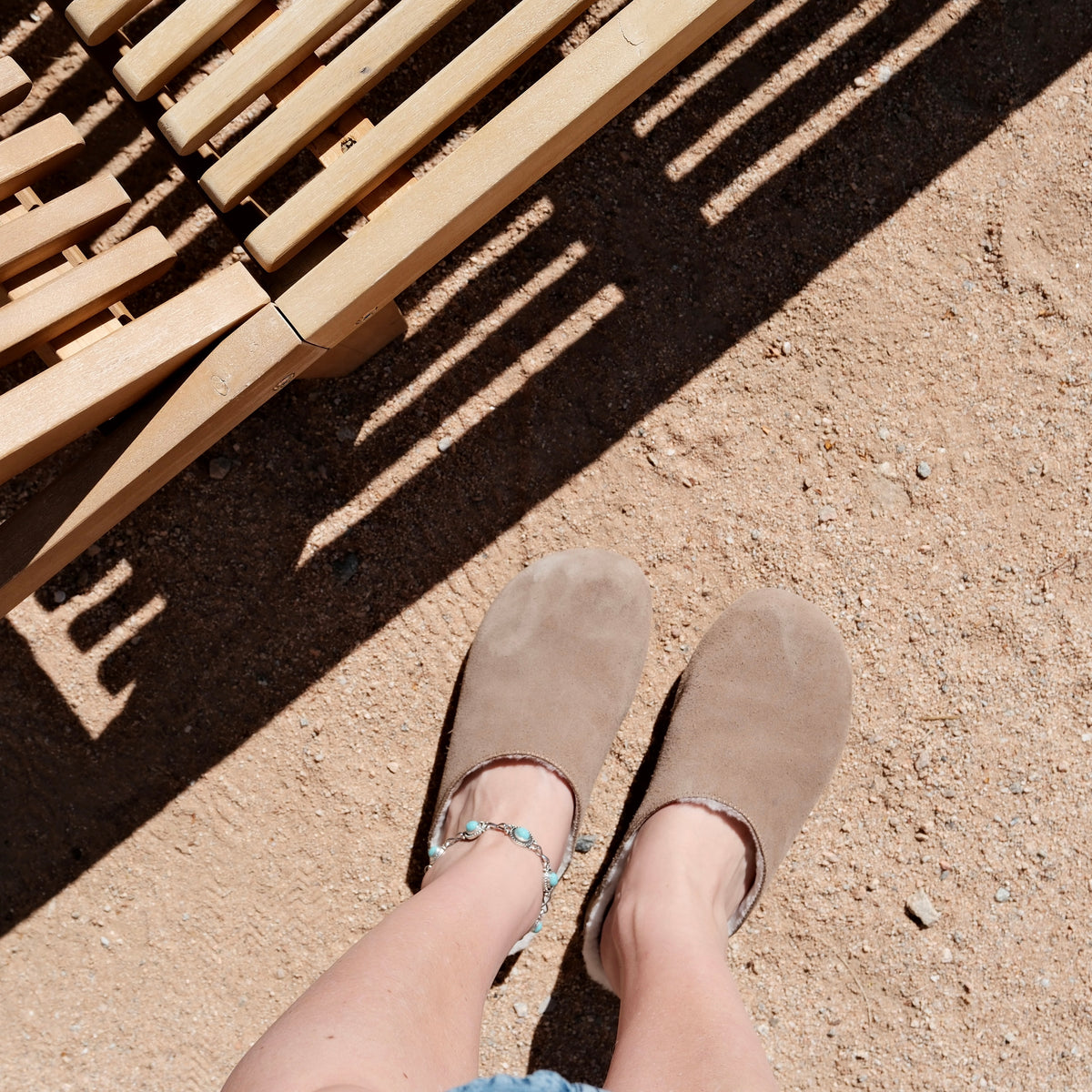 A top-down view of a person wearing brown suede clogs with turquoise embellishments on the straps, standing on sandy ground under the shadow of a wooden slatted structure.