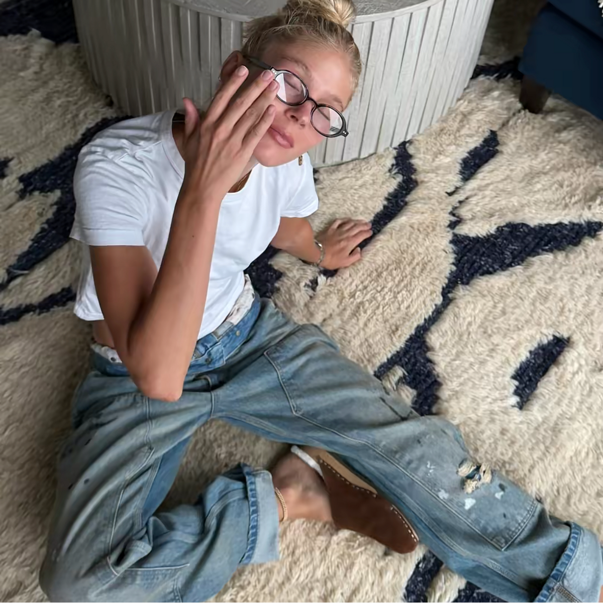 A young girl with blonde hair in a bun and glasses sits on a patterned cream and navy rug, wearing a white t-shirt, distressed light blue jeans and suede clogs. She is posing in front of a round ribbed white coffee table with a colorful game board on top.