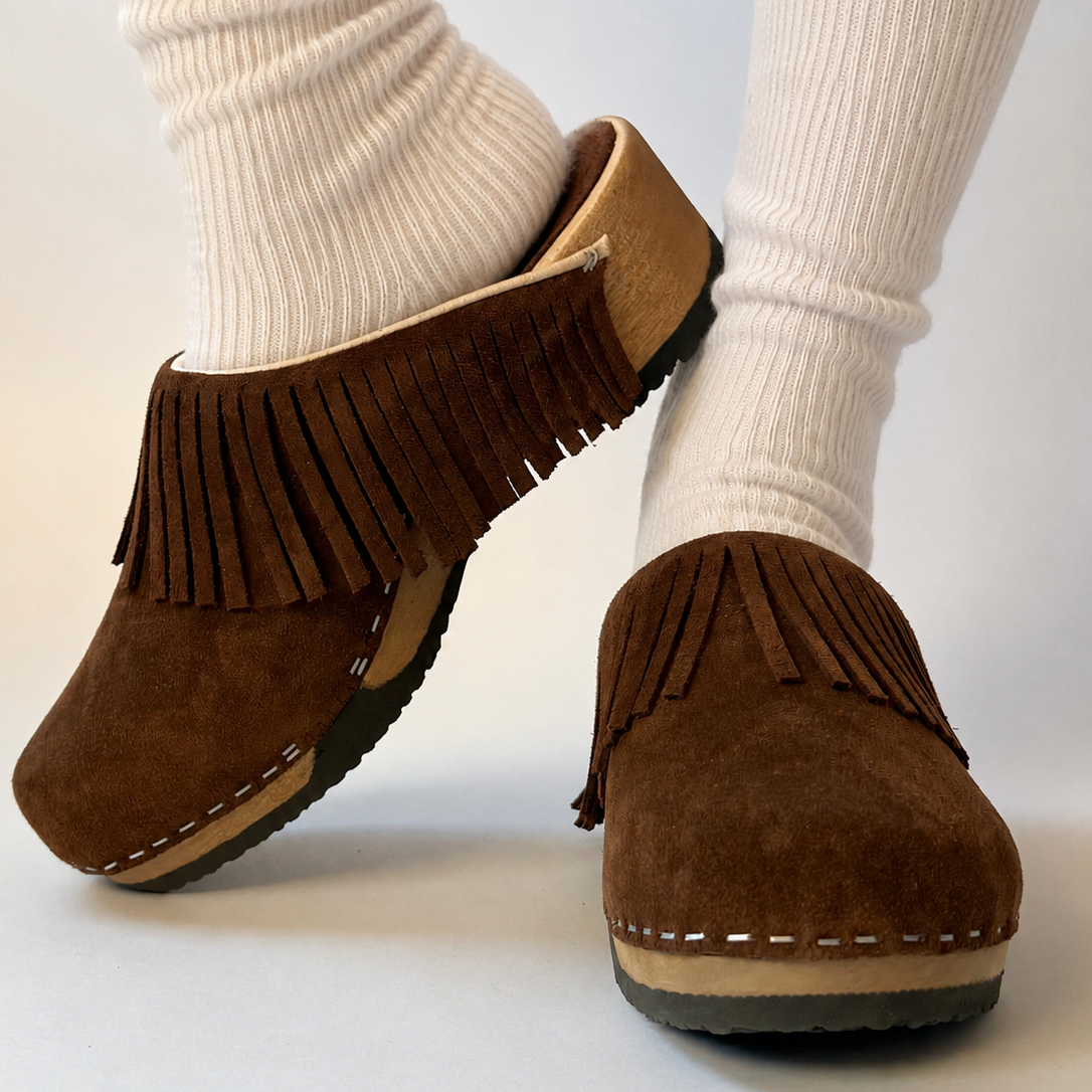 A pair of brown suede fringe mule clogs worn with off-white ribbed socks, shown against a plain light background.