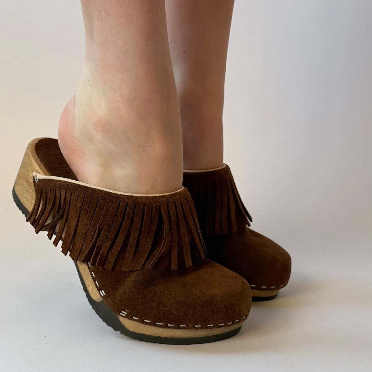 A pair of brown suede fringe wedge mule shoes worn on a person's feet, shown against a plain light gray studio background.