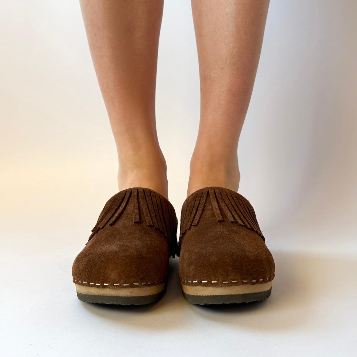 A pair of brown suede fringe mule shoes worn on a person's feet, shown from the front against a plain light background.