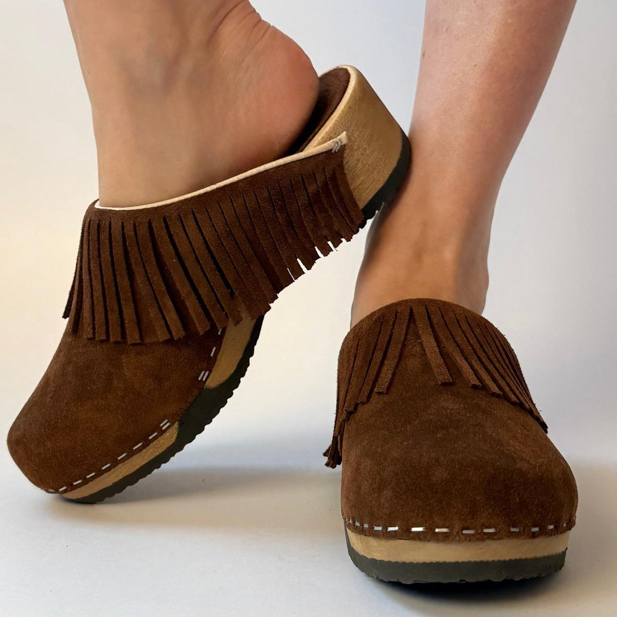 A pair of brown suede fringe mule clogs shown on a person's feet against a plain light background.