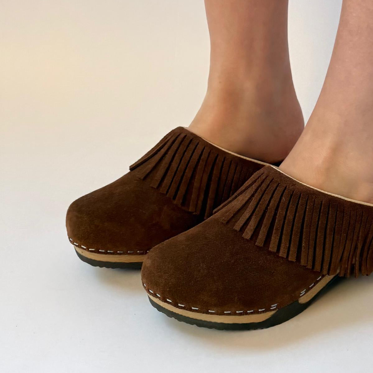 A pair of women's brown suede fringe mule shoes shown on a model's feet against a plain off-white background.