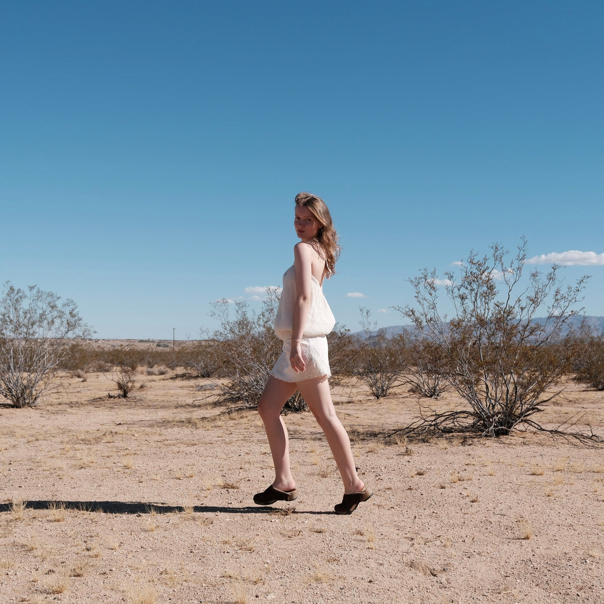 A woman wearing a light beige sleeveless dress and brown fringe clogs walks across a desert landscape under a clear blue sky.