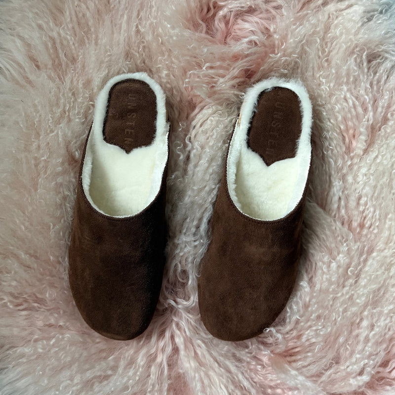 A pair of brown suede shearling clogs with a white faux-fur lining, displayed on a fluffy pink sheepskin rug.