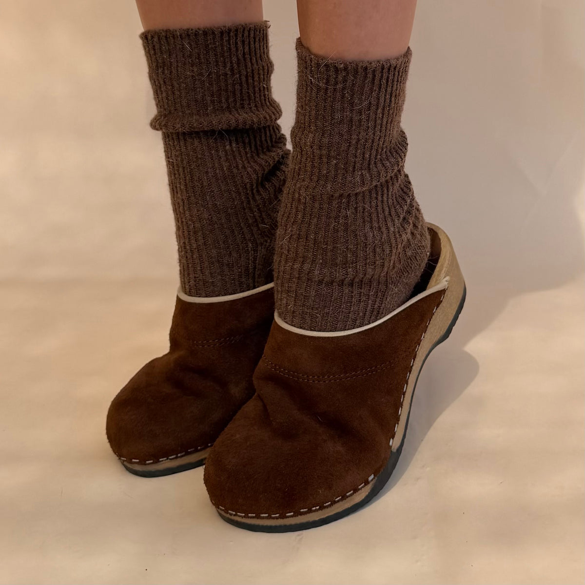 A person wearing brown knit socks and matching brown clogs with a light beige trim, standing against a plain light beige background.