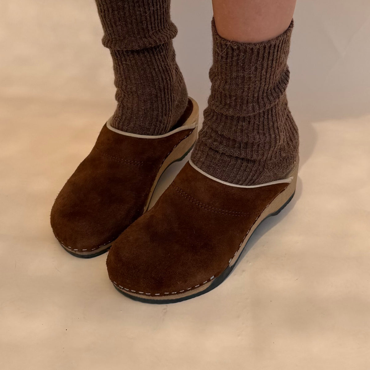 A pair of brown suede clogs with a white contrast trim, shown on a person's feet with brown ribbed socks against a neutral beige background.