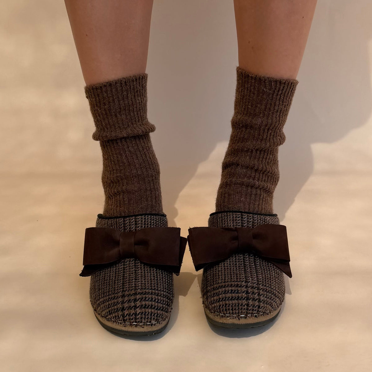 A person's legs wearing brown ribbed socks and matching brown knit clogs with large bow details on the front, photographed against a plain light background 