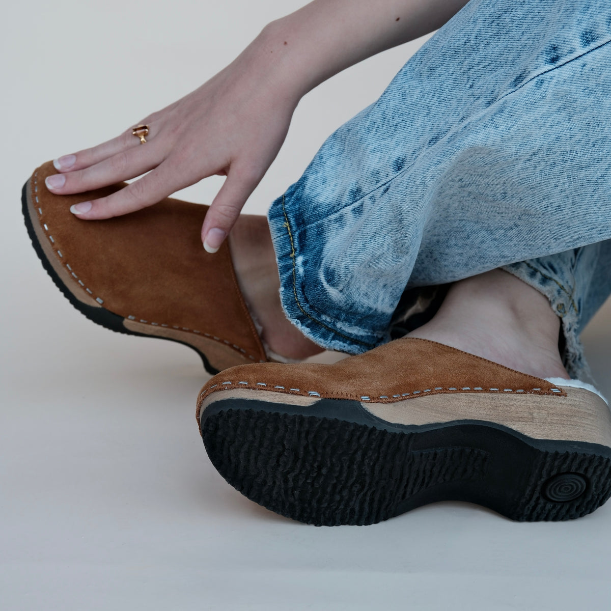a model sitting on a beige background, wearing brown clogs. 