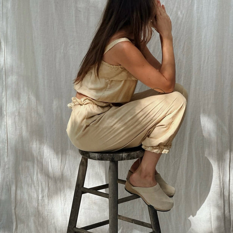 A woman wearing a beige linen two-piece set consisting of a sleeveless top, matching pants and a pair of KUNSTEN clogs, sitting on a dark wooden stool in front of a draped white fabric backdrop.