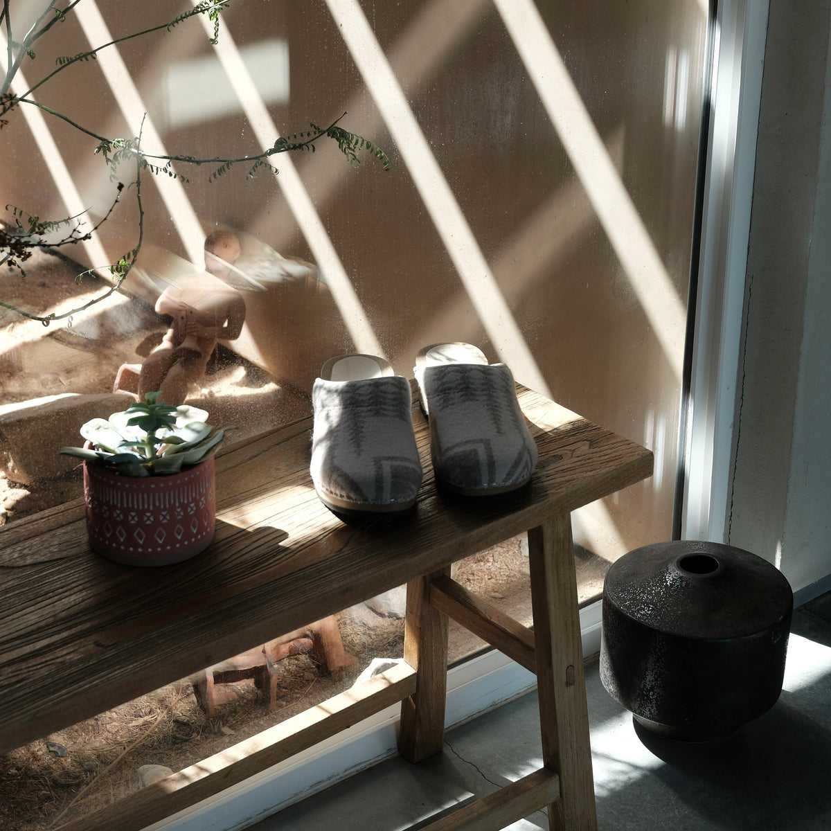 A pair of gray knit clogs with white geometric patterns, displayed on a rustic wooden bench in a sunlit room with dramatic window shadows.
