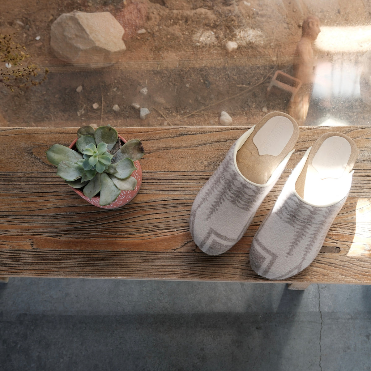 A pair of wool clogs in a light beige and gray pattern, placed on a rustic wooden table next to a small potted succulent.