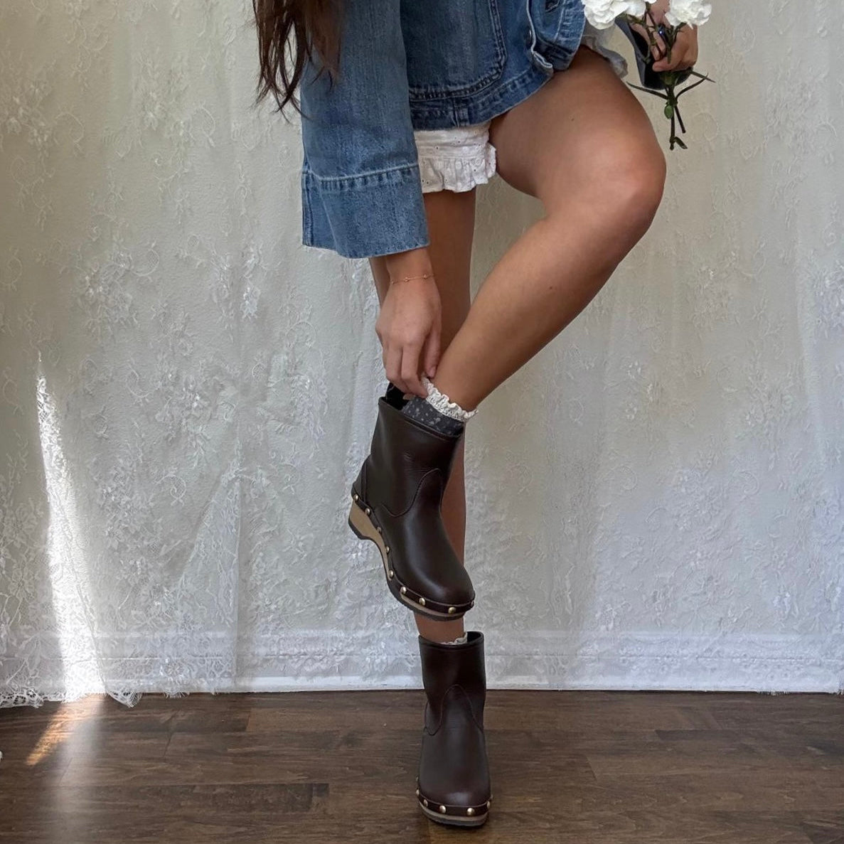 A person wearing a blue denim jacket and white ruffled shorts is shown from the waist down, adjusting a dark brown boot with metal studs while wearing the matching boot. They are holding a small bouquet of white flowers in front of a textured white fabric backdrop on a wooden floor.