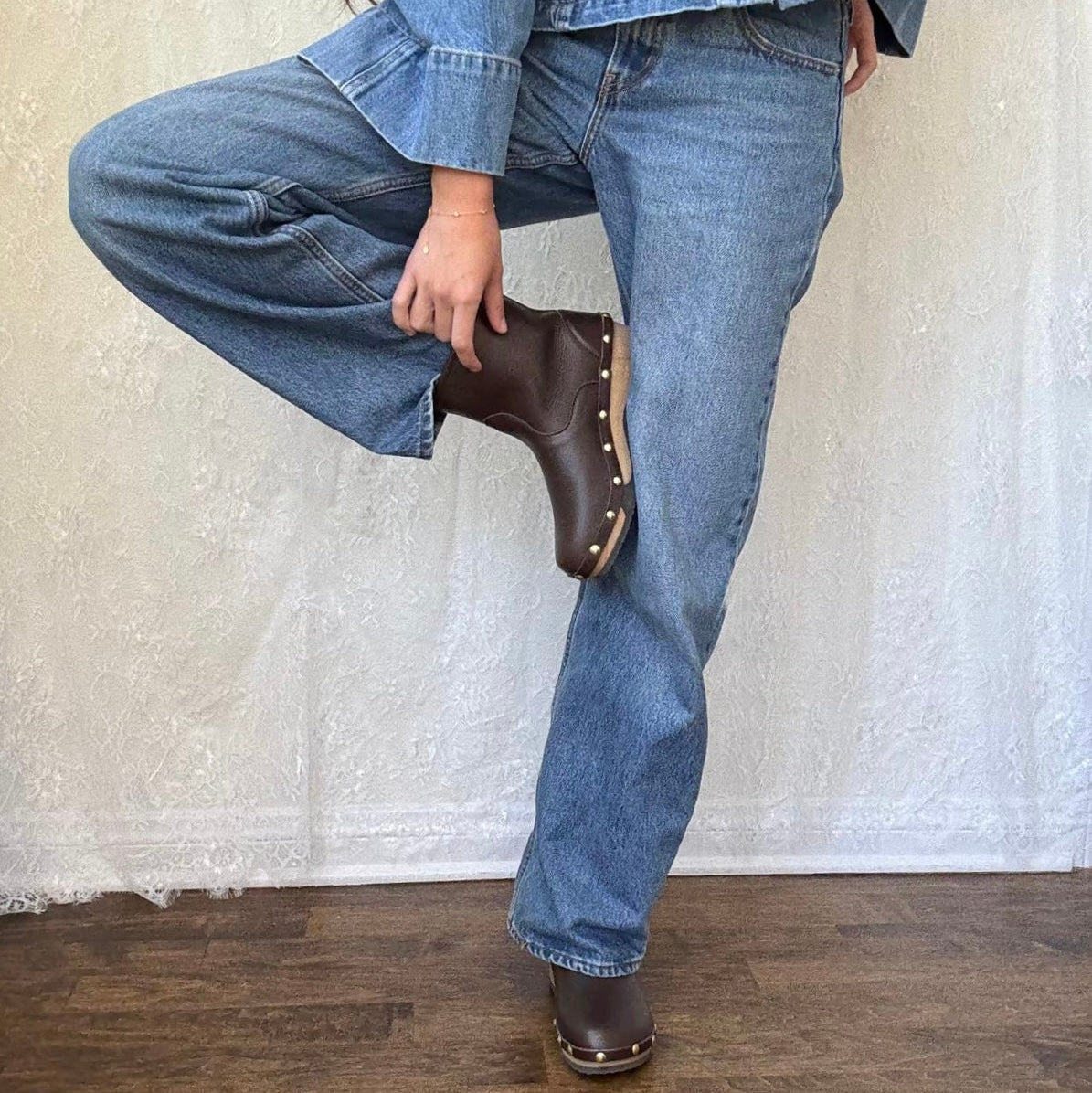 A person wearing blue denim jeans and a matching denim jacket, with one leg bent upward to show off a dark brown leather boot with gold-tone studs. The background is a textured off-white curtain with a dark wood floor.