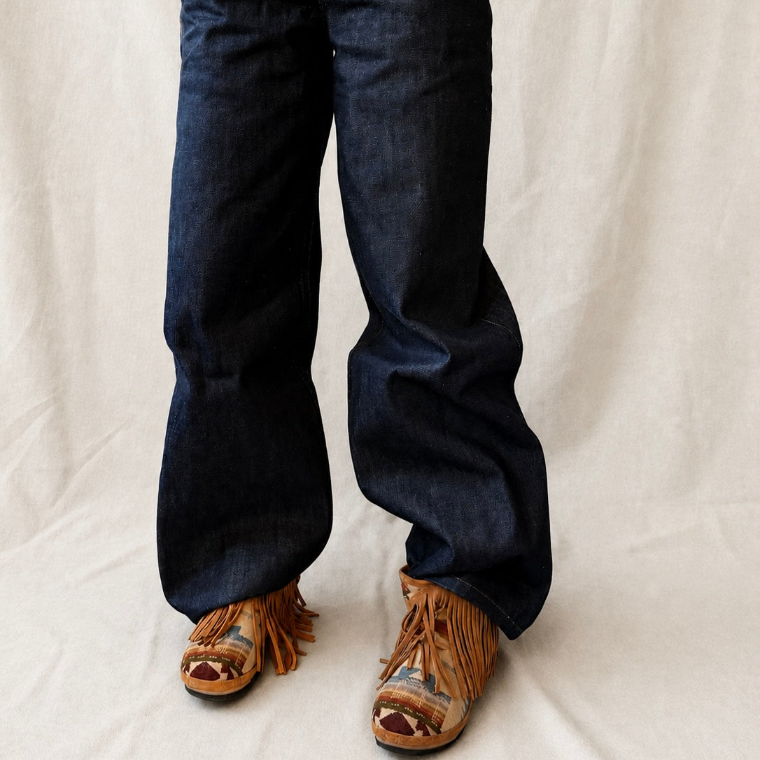 Close-up of a person's legs wearing dark blue wide-leg jeans and brown fringed clog boots against a draped off-white fabric backdrop.